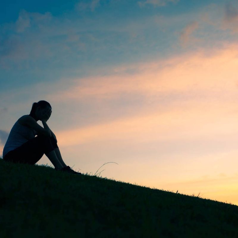 Silhouette of Woman Sitting on a Hill Depressed with Hands on Head Upset and Sad with Sunset Behind