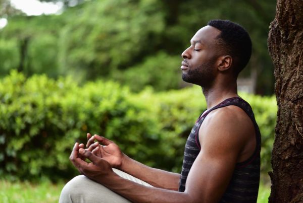 Man Meditating Next to Tree with Green Grass and Bush Behind Him