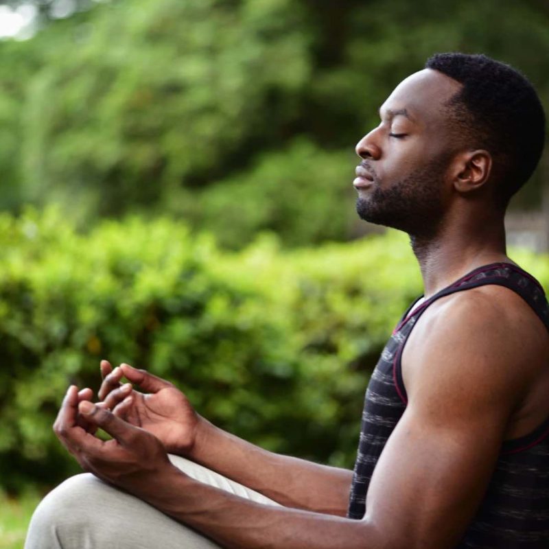 Man Meditating Next to Tree with Green Grass and Bush Behind Him