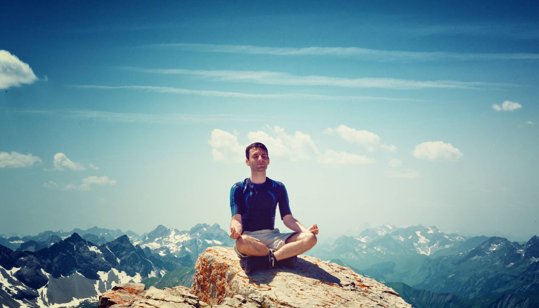 Man Meditating on Cliff with Blue Sky and Clouds in Background